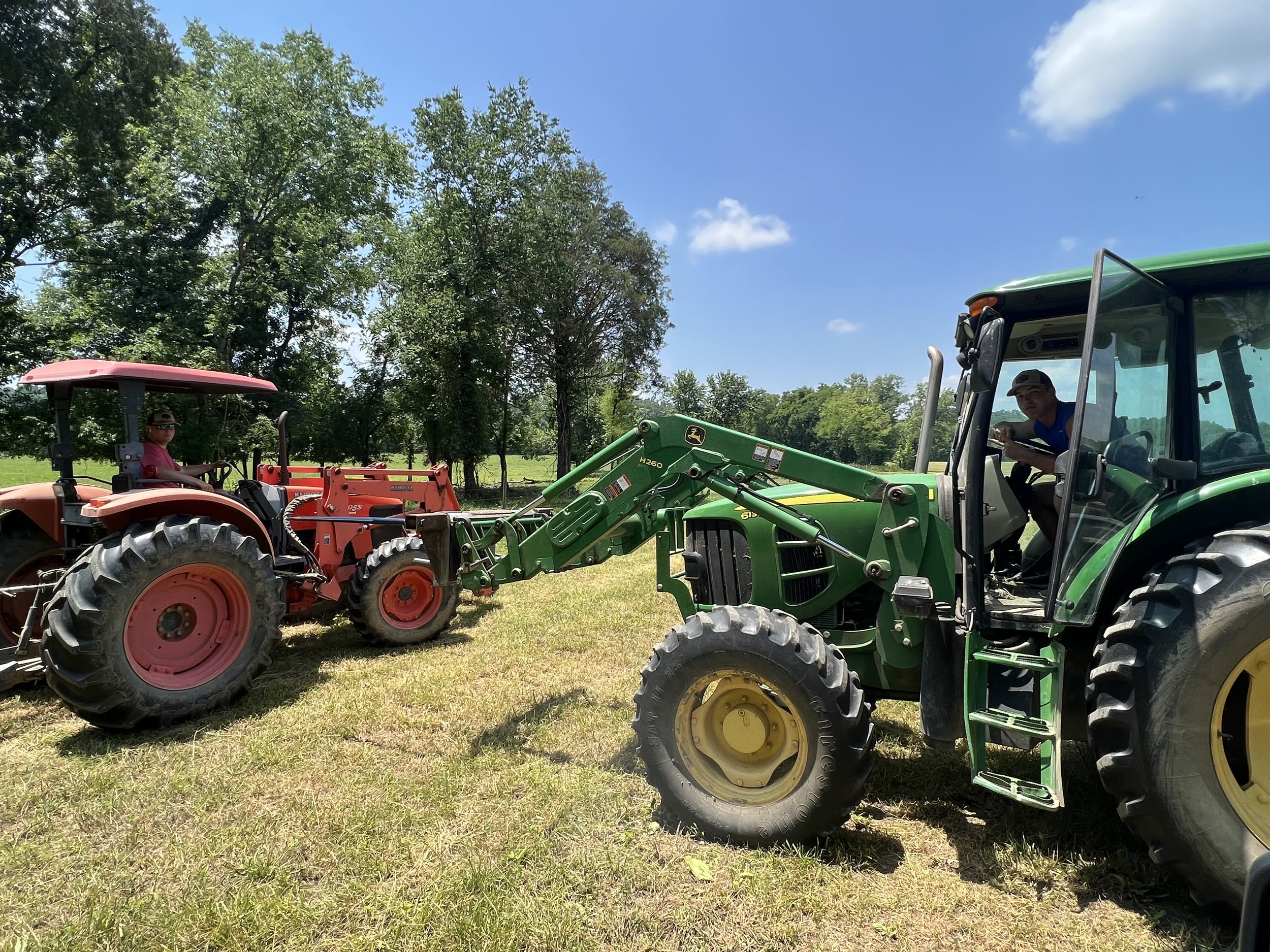Two tractors, one red and one green, are parked side by side on a sunny day in a grassy field. A person in a green tractor is operating the machinery while another individual is seated in the red tractor. Trees and a clear blue sky are visible in the background, emphasizing a rural farming setting.